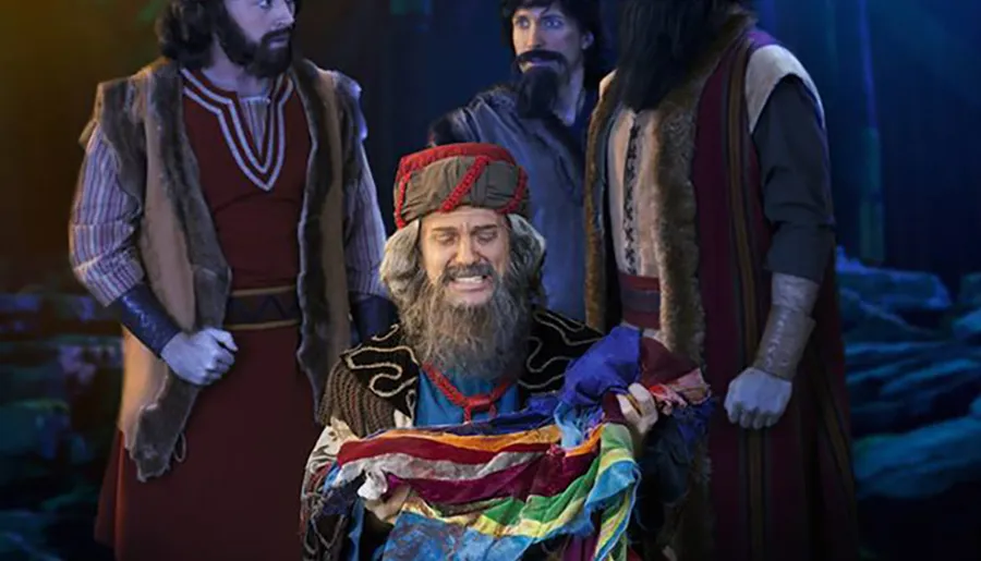 A person in a colorful costume appears to be emoting dramatically while holding a bundle of colorful fabric, surrounded by three other individuals in historical or fantasy attire, set against a dark, moody background suggestive of a theatrical or storybook setting.